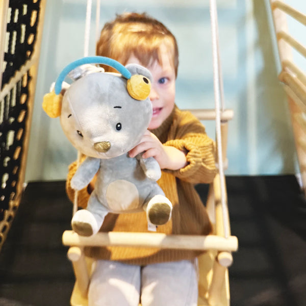 Kind sitzt auf einer Schaukel und hält Lenabie Aaron (Bär) – das Audio-Kuscheltier mit flauschigen Ohrenschonern als liebevoller Begleiter für Geschichten, Musik und Hörspiele.